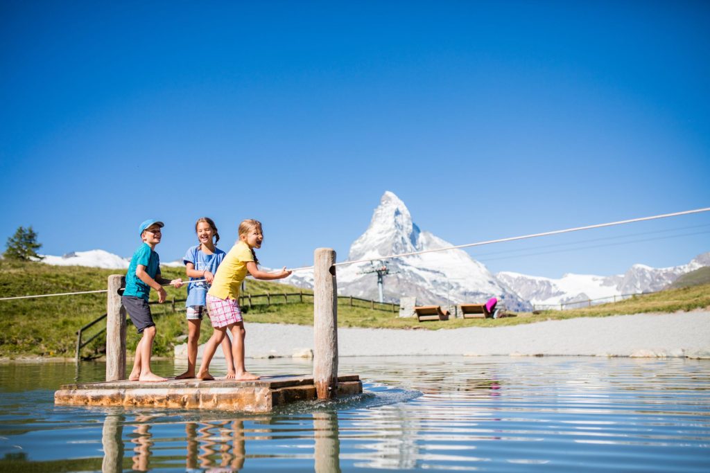 Children playing on a lake during their family summer holidays in the Alps, on a sunny day with the snowy Matterhorn in the distance.