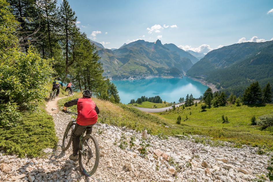 Mountain bikers on a broken stone path leading towards Tignes Lake, which can be seen in the distance - it's beautiful blue waters sitting in between mountains that tower above.