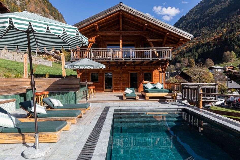 An outdoor swimming pool, sun loungers and parasols are found on the outdoor terrace in front of a wooden chalet, Chalet Doyen, with views to forest hills and blue skies seen in the background.