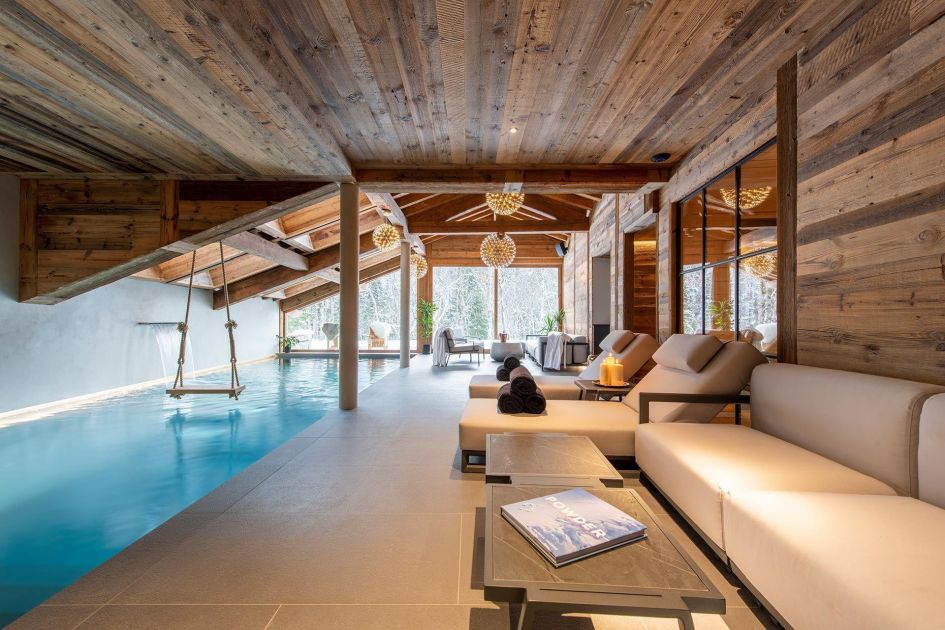 A large blue swimming pool sits to the left of the picture of Chalet Tataali's wellness SPA area, whilst lounge sofas are on the right of this image. A window wall at the far end of the room frames the views outside.