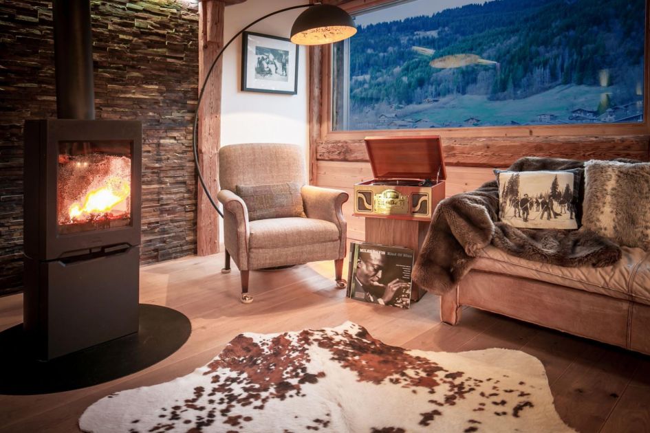 Chalet Vogue's interiors offer an inviting alpine setting. A cow rug, log burner, sofa, armchair and record player sit in the wooden setting before a landscape window with views to the forested hills outside.