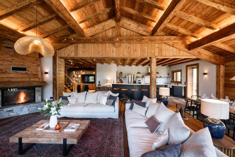 Vaulted wooden ceilings house Chalet Xanadu's homely living room. A cosy lounge area is in the foreground, adjacent to a wood-burning fireplace in the corner. At the back of the room, a modern open kitchen can be seen with a breakfast bar.