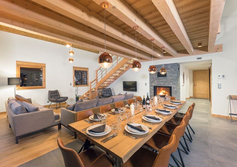 A long wooden dining table found in Colibri Penthouse's open living area. A lit wood-burning fireplace is in the background, alongside sofas and stairs leading to the second floor.