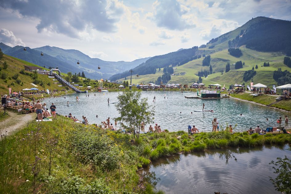 Picture of crowds enjoying all that this mountain lake has to offer, with some swimmers, paddleboarders and plenty of people sunbathing around the shoreline. A slide into the lake can be seen in the distance, as can an overhanging cable car and mountain views completing the backdrop.