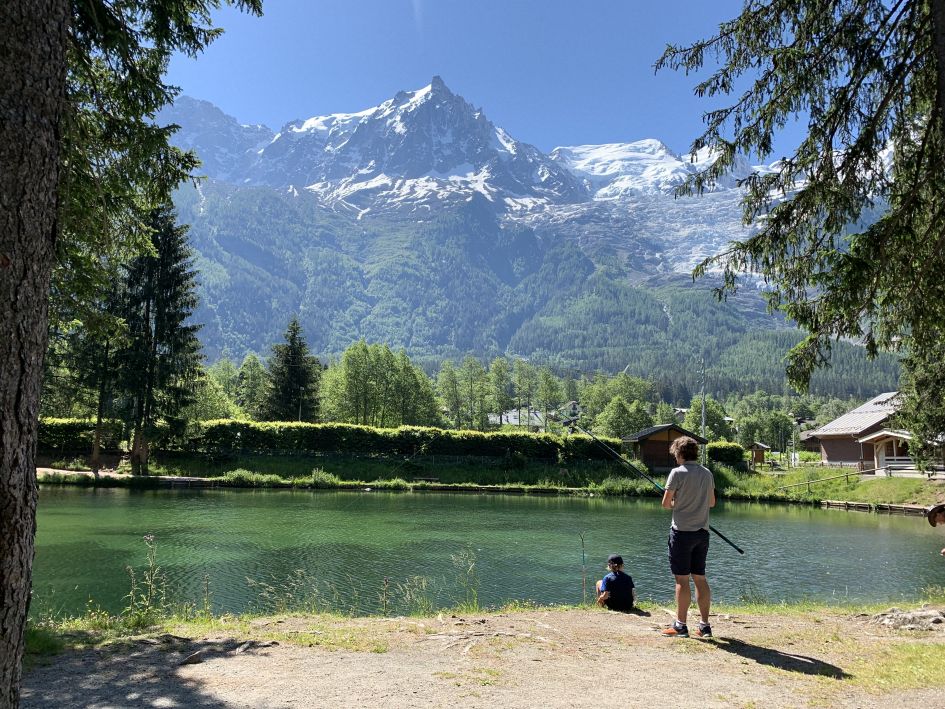 A man and his child fishing in an alpine lake that shimmers in the sun. An incredible snow-capped mountain can be seen in the distance, beyond the water and trees.