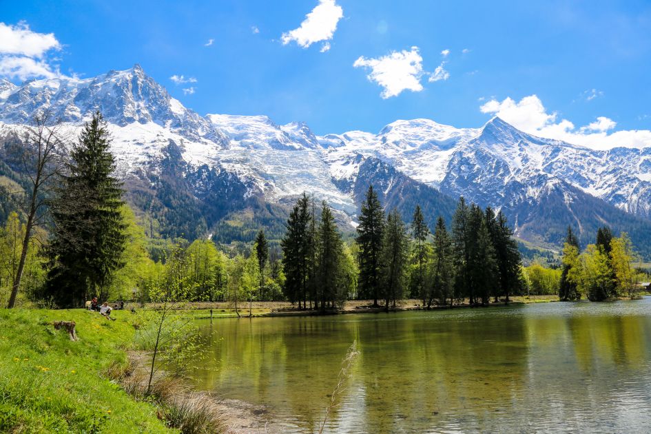 View of Lac des Gaillands in Chamonix, looking out to the water, green forest trees in the foreground and colossal snow-capped mountains in the background underneath blue skies - an excellent snapshot for the first picture featured in a summer holidays to alpine lakes blog!