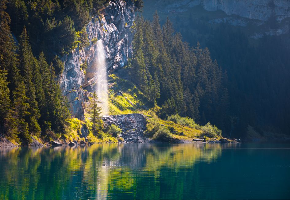 This waterfall to the side of a dark blue lake, Lake Oeschinen in Switzerland, shining in the light with trees surrounding, is an exceptional showcase for the natural beauty that summer holidays to alpine lakes provide visitors with!