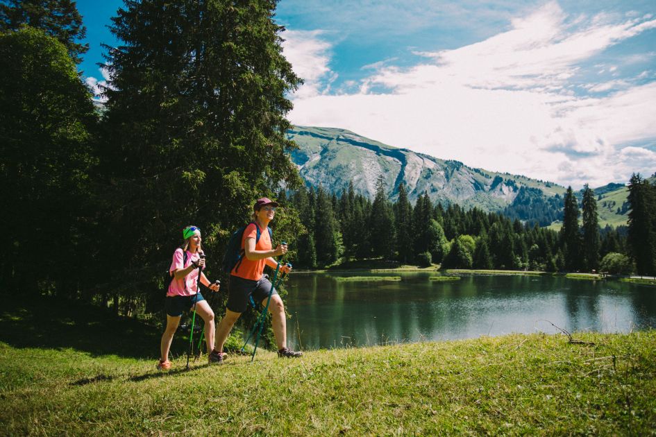 Two hikers with poles taking a stroll around a mountain lake, that's surrounded by coniferous green trees around the shoreline and mountain views in the background.