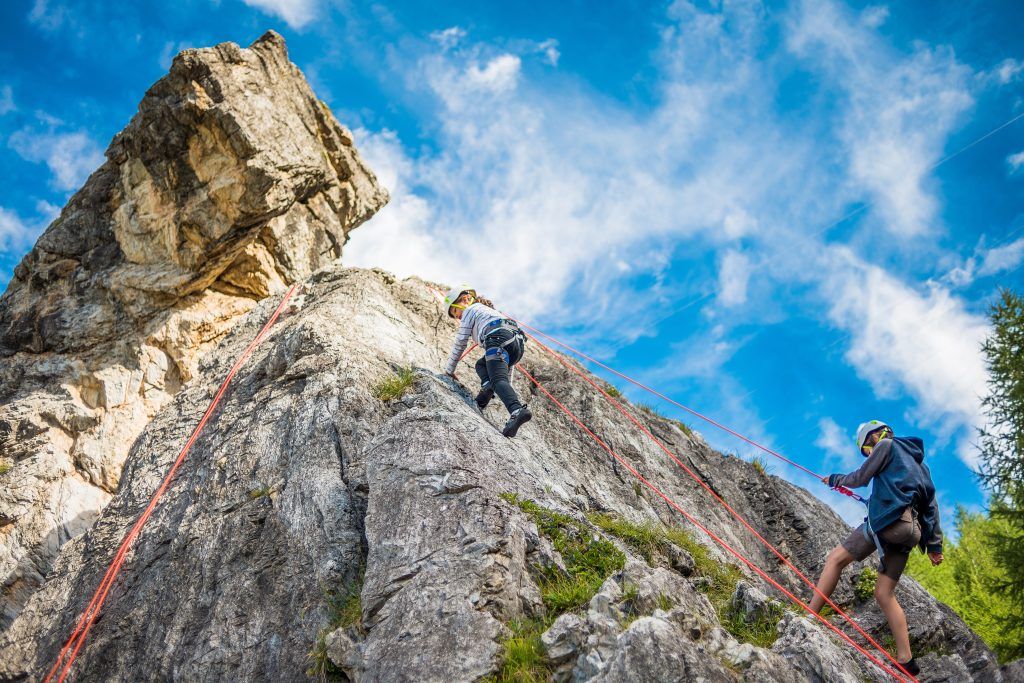 Children climbing on ropes up a craggy rock face with blue skies featuring. Photo specifically of rock climbing in Val d'Isère, one of the best family activities in the Alps.