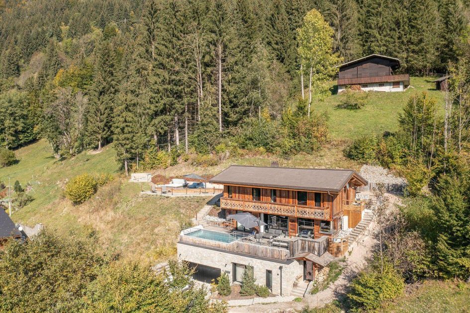 Exterior photo of Alpine Retreat, a wooden luxury summer chalet in Morzine, with a focus on the chalet's terrace which houses an outdoor swimming pool, surrounded by forestry in the background.