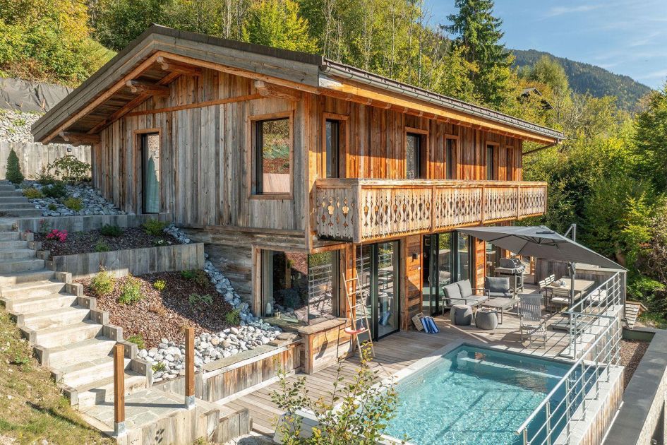 As one of the best summer chalets in Morzine, Alpine Retreat's stone and wooden exterior and outdoor terrace (featuring a swimming pool) can be seen shining in the sun, surrounded by forests in the background.