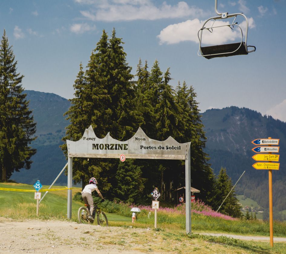 A mountain biker passes under a 'Morzine' sign to begin a mountain bike trail in Morzine bike park, with views of trees, the mountains in the background and chairlift above.