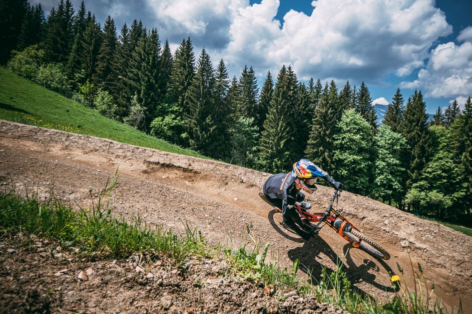 A mountain biker speeds down a turn in a downhill dirt track, in front of green trees and cloudy blue skies.