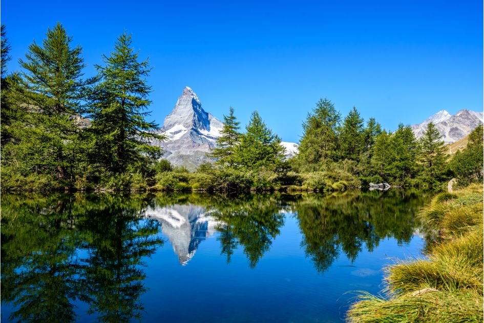 Lake Grindjisee sits beneath blue skies and is surrounded by trees, whilst the iconic Matterhorn Mountain is reflected in the water - perfect for luxury walking holidays in Europe, and for a lakes and mountains holiday in the Alps!