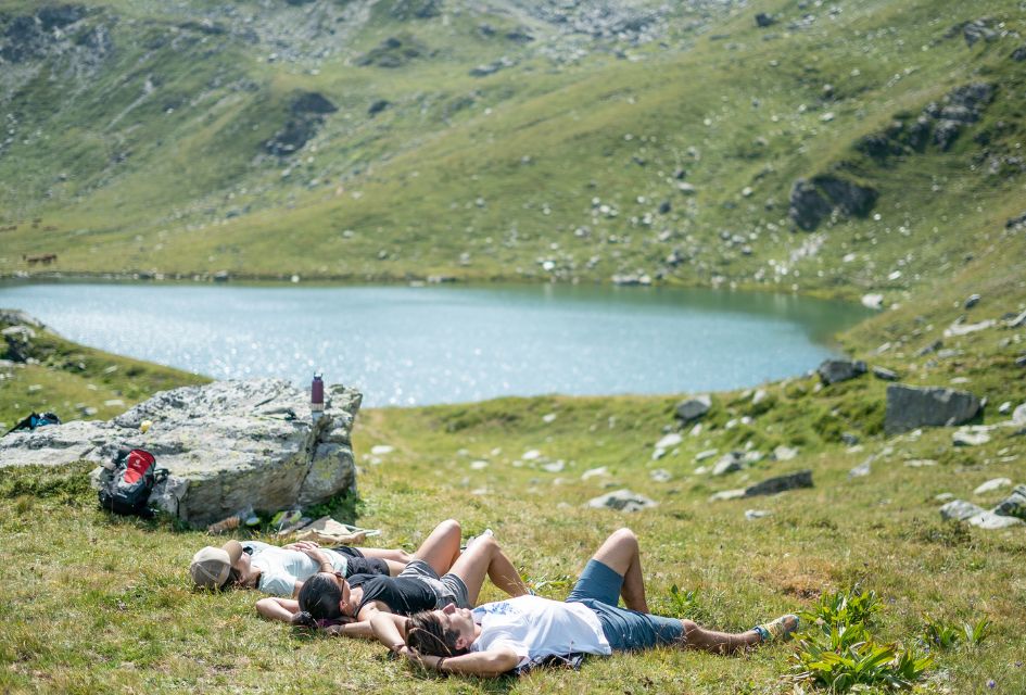 Three friends laying down to relax on the grassy mountains of St Martin de Belleville in summer after a hike, with a sparkling lake in the background.