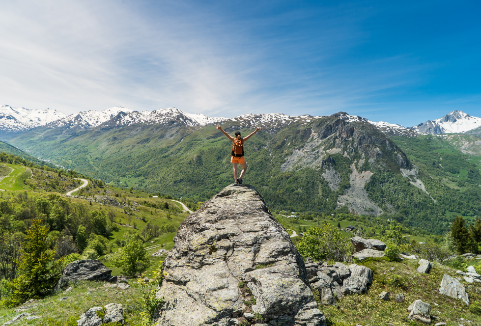 Woman with backpack standing on a rock with arms out, celebrating her hiking view of mountains, with snowy peaks in the distance.