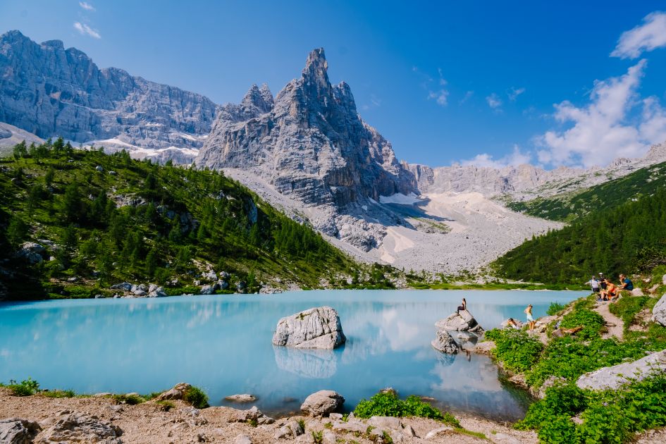 Bright turquoise water of Lago di Sorapis, with mountain peaks, lush trees, and blue skies surrounding this beautiful mountain lake in the Dolomites.