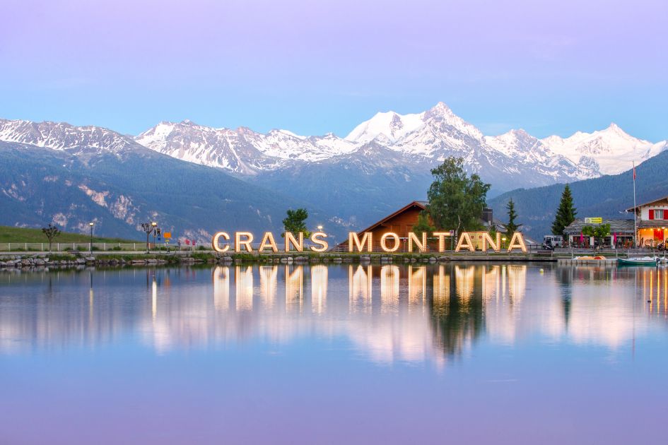 Photo of Lake Grenon with the 'Crans Montana' sign reflected in its waters and mountains in the background.