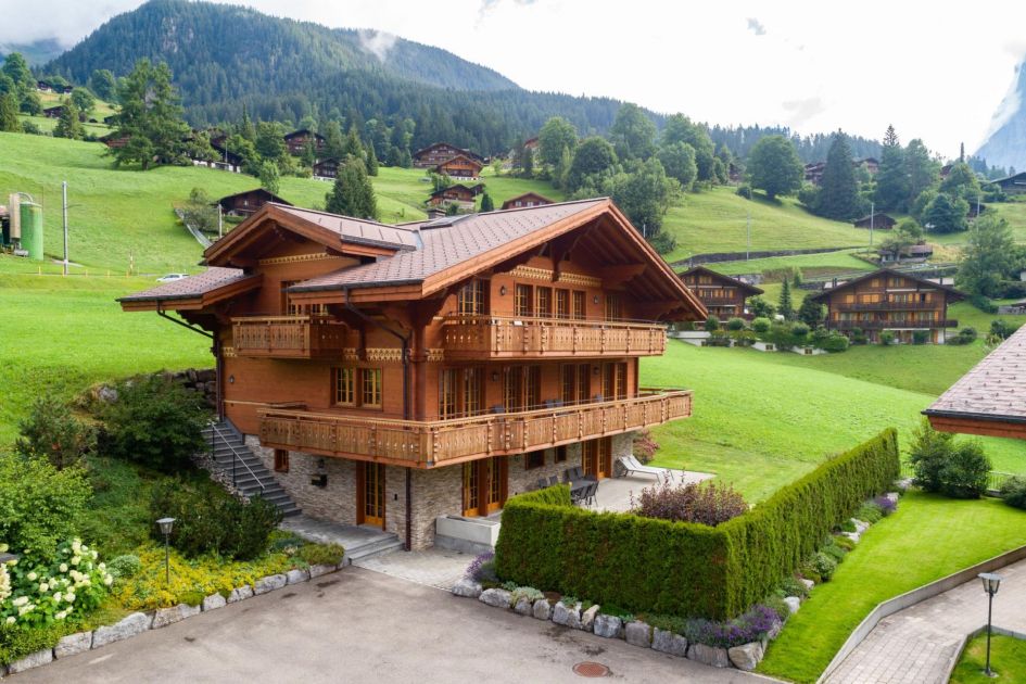 Wooden and stone exterior of Chalet Alia in Grindelwald, surrounded by greenery and the mountains of Switzerland in the background.