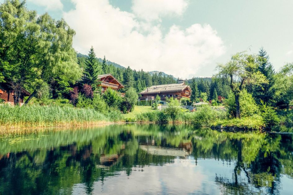 Exterior of UItima Crans Montana's two chalets hidden amongst the trees, behind the pond that features on its grounds. This property is just a minute's walk from Chermignon Lake perfect for exporing the lakes and mountains in Switzerland.