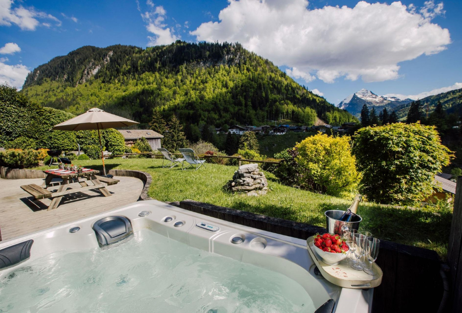 Hot tub outside Chalet Bizet, with mountain views and a tray with champagne and strawberries on the side. Also looking over the garden and the picnic table on the decking.